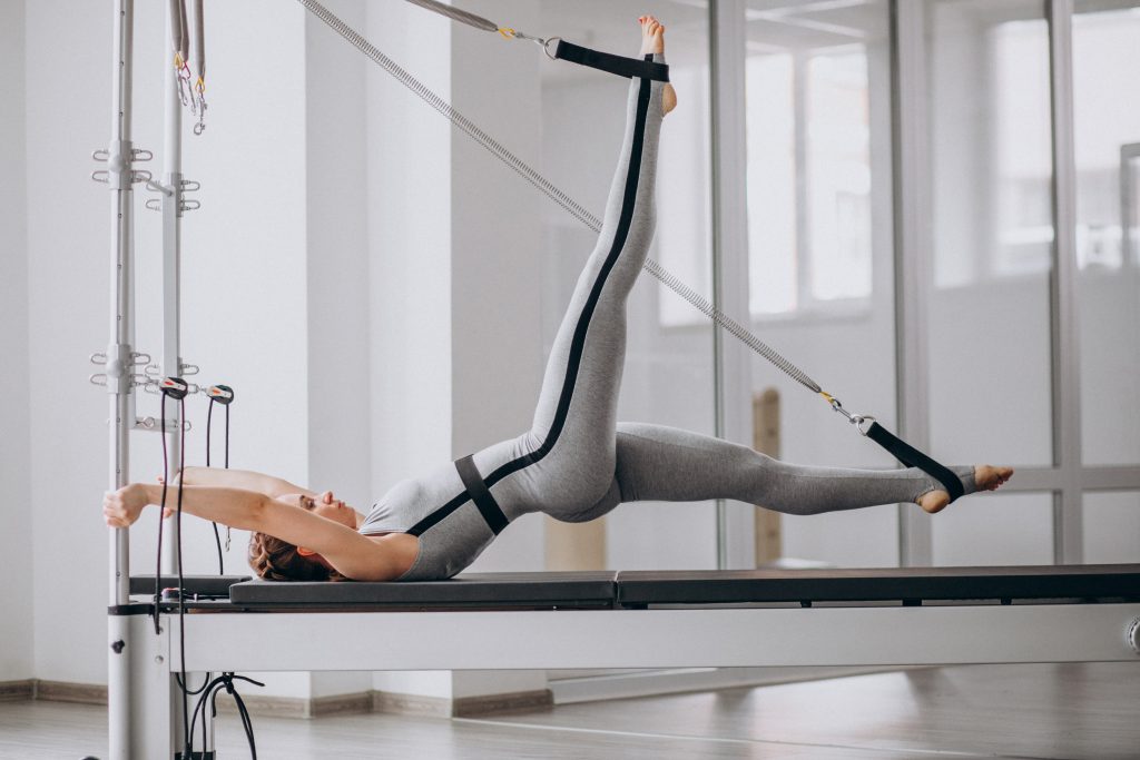 Persona sobre una máquina de Pilates reformer apoyada sobre la cama boca arriba y sus pies enganchados en las poleas haciendo un ejercicio 